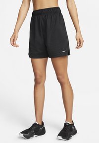 Femme portant un short de sport noir Nike et des chaussures de course noires Nike, debout avec les bras détendus le long du corps sur fond blanc.