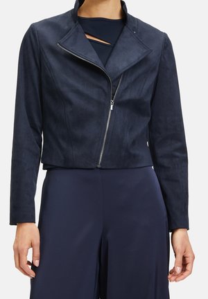 Femme portant une veste courte bleu marine avec une fermeture éclair asymétrique et un pantalon large bleu marine, montrant le haut du corps et les mains.
