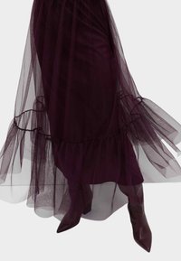 Płynąca fioletowa spódnica z tiulu z marszczonym dołem, zestawiona z bordowymi botkami na klockowym obcasie z migdałowym noskiem, na białym tle.
