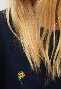 Suéter azul marino con una flor amarilla bordada en el pecho, con detalles de tallo verde. El cabello rubio largo y liso obstruye parcialmente el diseño.