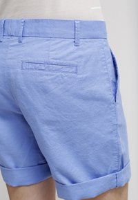 Shorts en coton bleu avec un ourlet replié, dotés de deux poches latérales et d'une seule poche arrière. Texture lisse et tissu léger.