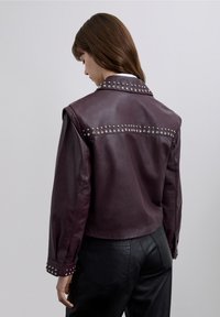 Veste en cuir bordeaux avec des clous en argent le long des épaules et des poignets, design court et texture lisse et brillante.