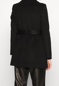 Cappotto di lana nera con un design su misura, caratterizzato da una vita cintrata e maniche lunghe. Tessuto liscio, taglio dritto e senza motivi visibili.