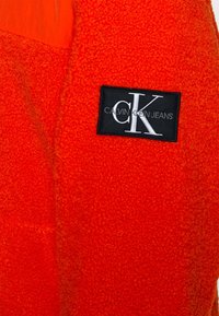 Punainen fleecepaita, jossa on musta etiketti valkoisella tekstillä: "CALVIN KLEIN JEANS". Esittelee pehmeän tekstuurin ja elävän värikontrastin.