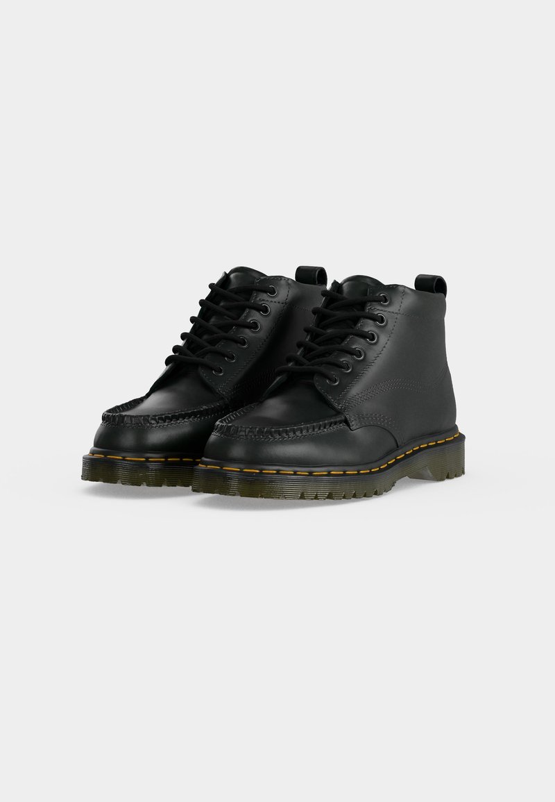 Paar schwarze Leder-Stiefeletten mit dicker Sohle, gelber Naht und schwarzen Schnürsenkeln auf hellem Hintergrund.