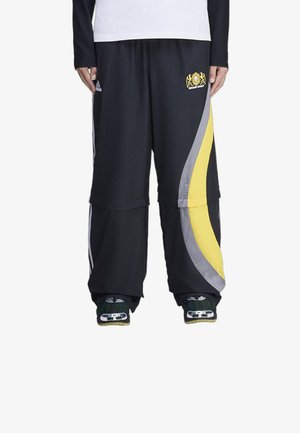 Persona vistiendo pantalones negros de pierna ancha con rayas curvas amarillas y grises, logo de Adidas y zapatillas deportivas verdes y blancas.