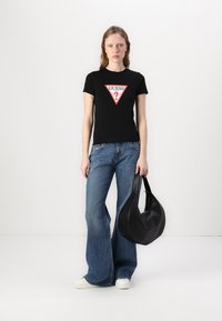 T-shirt noir à manches courtes avec un logo rouge en forme de triangle "GUESS?", associé à un jean bleu évasé et un sac à main noir de style hobo.