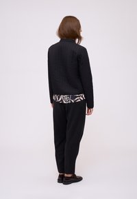 Giacca nera trapuntata con maniche lunghe, indossata sopra una maglietta blu stampata. I pantaloni neri, le scarpe marroni e uno sfondo neutro completano il look.