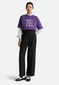 Paarse t-shirt met de tekst "Ken je Waarde", gedragen over een witte blouse. Zwarte hoge taillebroek, zwarte flats en grijze sokken.