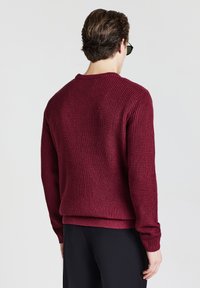 Mann, der einen strukturierten burgunderfarbenen Pullover und schwarze Hosen trägt, von hinten vor einem schlichten hellen Hintergrund betrachtet.