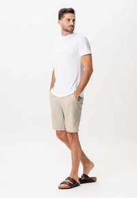 Mand står med hænderne i lommerne på sine beige shorts, iført en simpel hvid t-shirt og brune sandaler med to stropper, og kigger mod venstre.