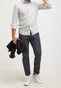 Camicia a righe bianche e grigie con bottoni, pantaloni blu, giacca nera e sneakers bianche. La camicia ha le maniche arrotolate e bottoni ben visibili.