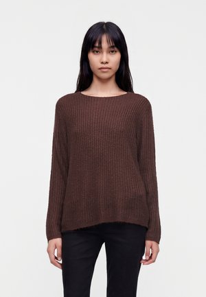 Jeune femme portant un pull marron texturé à manches longues et un pantalon noir, debout devant un fond blanc uni.