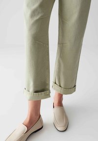 Pantalons vert clair à revers, avec des motifs de couture visibles et une finition texturée, associés à des chaussures beiges à enfiler.