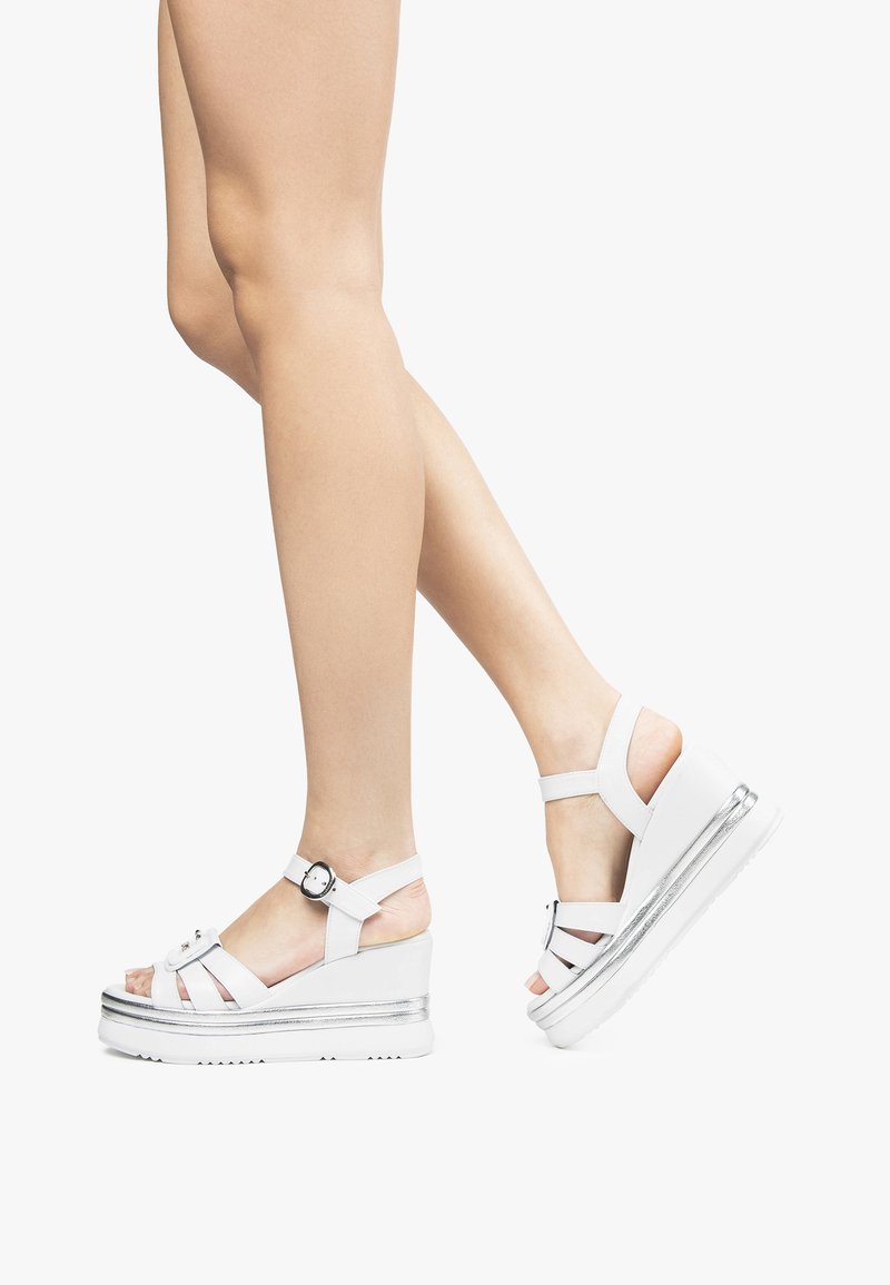 Sandalias de plataforma blancas con un acento plateado, con múltiples correas y cierre de hebilla en el tobillo. Textura suave y diseño de talón redondeado.