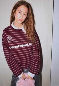 Bordó csíkos hosszú ujjú ing fehér logóval, alatta fehér galléros ing; a modell egy rózsaszín bolyhos táskát tart.