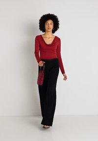 Rdeča majica z dolgimi rokavi in drapirano ovratnikom, v kombinaciji s širokimi črnimi hlačami. Model drži majhno rdečo perlirovo torbico.