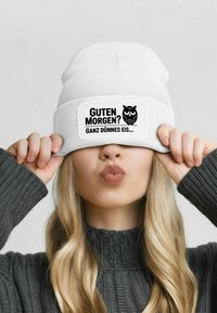 White knit beanie with a black label displaying "Guten Morgen?" and an owl design. Worn low, covering part of the face.