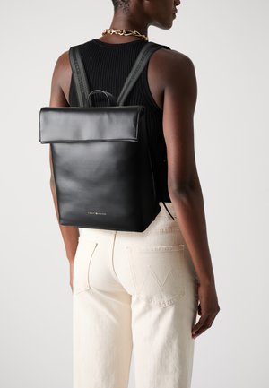 Sac à dos en cuir noir avec une fermeture à rabat, de forme rectangulaire, des bretelles ajustables et un logo métallique en bas. Texture lisse.