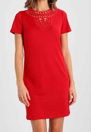 Czerwona sukienka z krótkim rękawem, z ozdobnym dekoltem przypominającym szydełkowanie, noszona przez osobę stojącą na jednolitym tle.