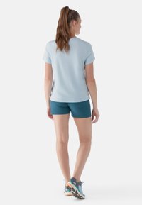 Top atletico a maniche corte di colore azzurro chiaro abbinato a pantaloni corti color teal, che mostra una vestibilità rilassata e una superficie morbida. Modella che indossa scarpe da corsa multicolori.