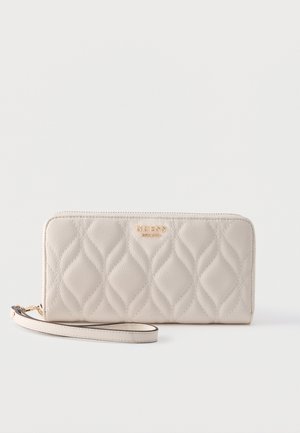 Kremowy pikowany portfel na nadgarstek z złotym logo „GUESS” i zamkiem błyskawicznym na płaskiej, białej powierzchni.