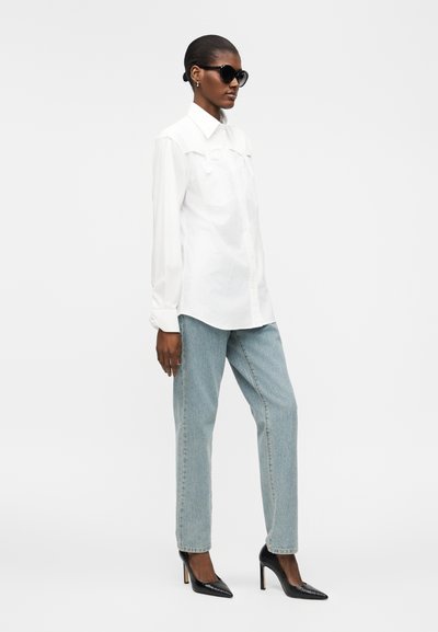 Femme portant une chemise blanche oversize, un jean bleu clair, des talons hauts noirs et des lunettes de soleil noires, debout devant un fond blanc uni.