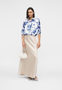 Λευκό και μπλε πουκάμισο με κουμπιά και μοτίβο, μπεζ μακριά φούστα, λευκή τσάντα με πτυχώσεις και ανοιχτόχρωμα μυτερά παπούτσια. Λιτές υφές, άνετη γραμμή.