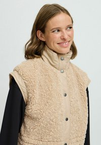 Beige ärmellose Weste mit einer flauschigen Textur, gestepptem unteren Abschnitt und Druckknopfverschluss. Sie hat einen hohen Kragen und eine lockere Passform.