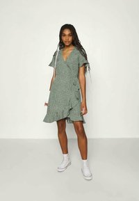 Vestido verde com bolinhas brancas, mangas curtas com babados e bainha assimétrica, combinado com ténis brancos. Textura do tecido suave.