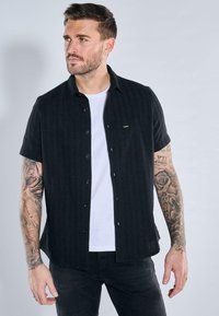 Kurzärmliges, knöpfbares Hemd aus schwarzem, strukturiertem Stoff mit einem dezenten Streifenmuster. Trägt ein weißes T-Shirt darunter und schwarze Jeans.