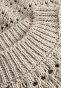 Beež kudumiga kampsun, millel on tekstureeritud soonikuga krae, avatud silmuste detailid ja jäme lõng, rõhutades pehmust ja soojust.