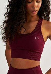 Bordowy sportowy crop top z teksturowanymi panelami, okrągłym dekoltem i małym logo serca po lewej stronie. Lekka, dopasowana konstrukcja.