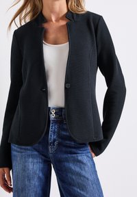 Blazer noir texturé avec un tissage léger, doté d'un col montant, d'une fermeture à boutons sur le devant, porté avec un jean en denim bleu.