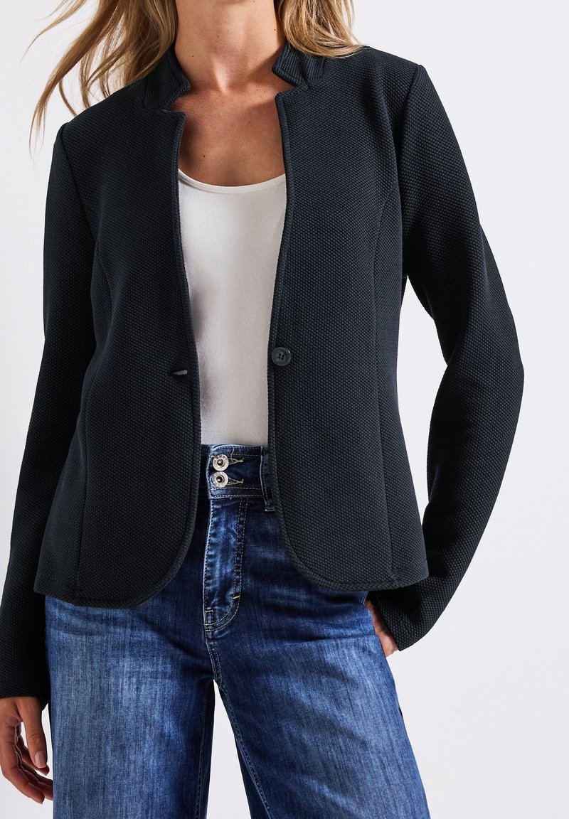 Blazer noir texturé avec un tissage léger, doté d'un col montant, d'une fermeture à boutons sur le devant, porté avec un jean en denim bleu.