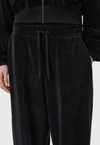 Svarta velour byxor med dragsko i midjan, sidofickor och subtil branding på benet. Mjuk textur och avslappnad passform.