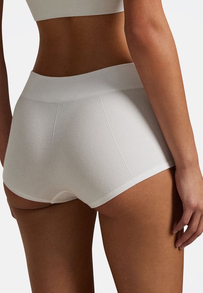 Kobieta ubrana w białą, wysoką, bezszwową bieliznę prążkowaną oraz dopasowany krótki top, przedstawiona od tyłu na jednolitym tle.