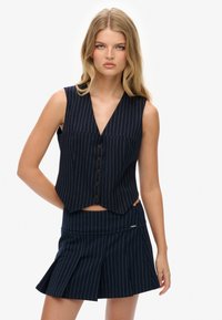 Granatowa kamizelka w prążki z głębokim dekoltem w kształcie litery V i pięcioma brązowymi guzikami, zestawiona z dopasowaną mini spódnicą w prążki z fałdami.