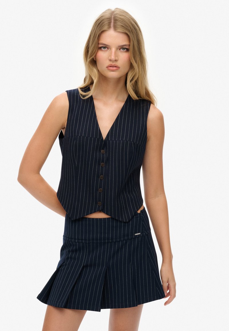 Granatowa kamizelka w prążki z głębokim dekoltem w kształcie litery V i pięcioma brązowymi guzikami, zestawiona z dopasowaną mini spódnicą w prążki z fałdami.