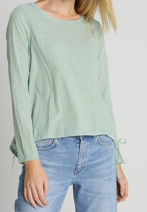 Femme portant un haut côtelé vert clair à manches longues avec des liens sur les côtés, associé à un jean taille haute bleu clair sur un fond uni.