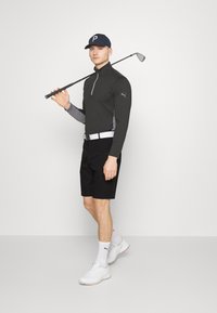 Czarne sportowe szorty, szaro-czarny longsleeve z zamkiem, biały pasek, białe trampki i granatowa czapka. Trzyma kij golfowy.