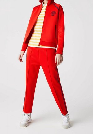 Osoba ubrana w czerwoną kurtkę na zamek i pasujące czerwone spodnie z granatowymi paskami po bokach, żółto-białą koszulkę w paski oraz białe trampki.