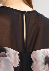 Černá průsvitná blůza s květinovým potiskem na spodní části, s knoflíkovým zapínáním na zádech a jemným rozparkem pro vrstvení.