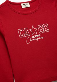 Czerwona, prążkowana koszula z długim rękawem z białym napisem graficznym "CA 82" oraz "Enjoyful Champion" z akcentem w postaci gwiazdki. Wykonana z miękkiego materiału.