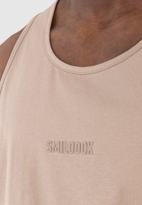 Läheltä kuvattu henkilö, jolla on yllään vaaleanbeige hihaton paita, jossa on rinnassa kohotetuin kirjaimin sana "SMILODOX".