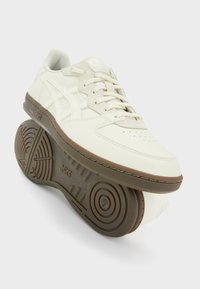 Bílá teniska s hladkým koženým svrškem, béžovými akcenty, perforovanou špičkou a gumovou podrážkou s kruhovými vzory na běhounu.