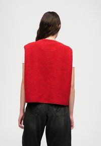 Sarkans vilnas veste ar teksturētu, pūkainu virsmu, plašiem izgriezumiem rokām un apaļu kakla izgriezumu, uzvilkta virs baltas krekla un melnām džinsām.