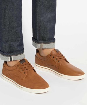Personne portant des baskets basses en cuir marron avec des semelles blanches et un jean bleu foncé retroussé sur un fond clair.