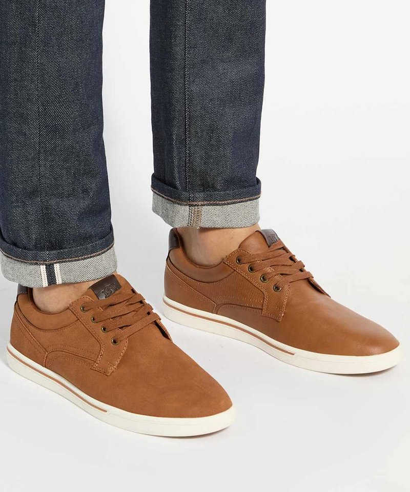 Personne portant des baskets basses en cuir marron avec des semelles blanches et un jean bleu foncé retroussé sur un fond clair.