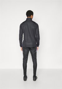 Zwarte sportjack met hoge kraag en lange mouwen, gecombineerd met zwarte broek met witte zijstrepen. Gladde, synthetische stof.
